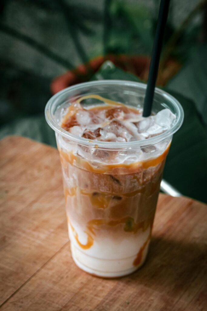 Close-up of iced caramel macchiato with straw on wooden table, surrounded by plants.
