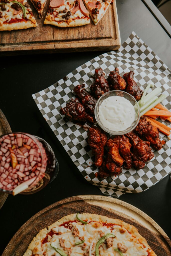 Overhead view of assorted pizzas and BBQ chicken wings with dipping sauce.