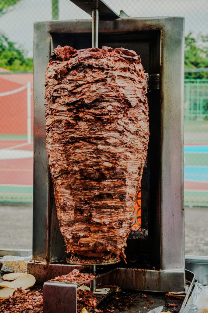 Juicy al pastor meat rotating on a spit, perfect for tacos, outdoors at a food stall.