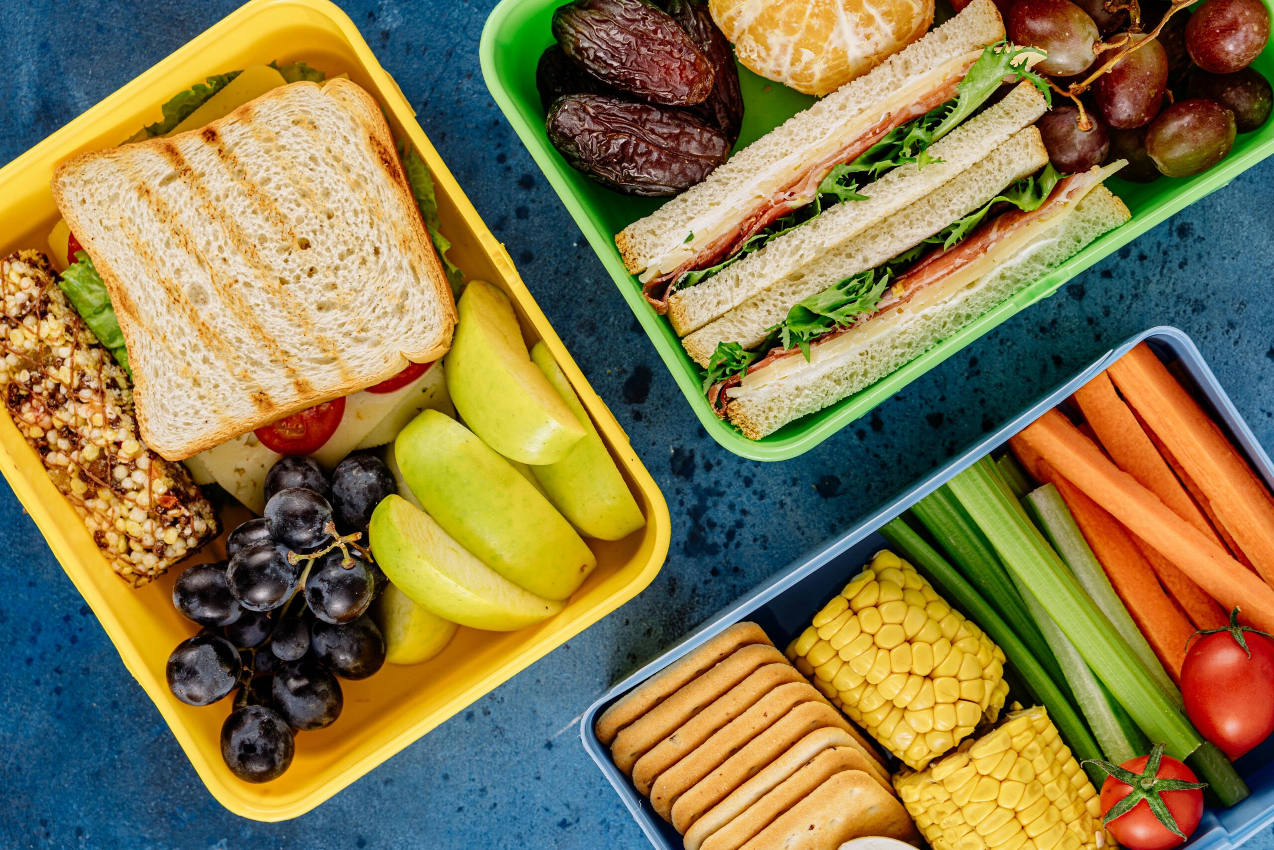 An array of healthy lunch box items with sandwiches, fruits, and vegetables on a blue background.