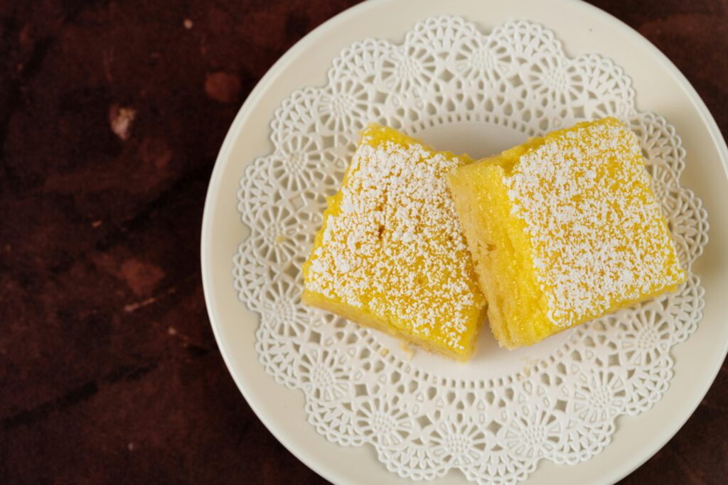 Two lemon bars on a doily-covered plate, dusted with powdered sugar.