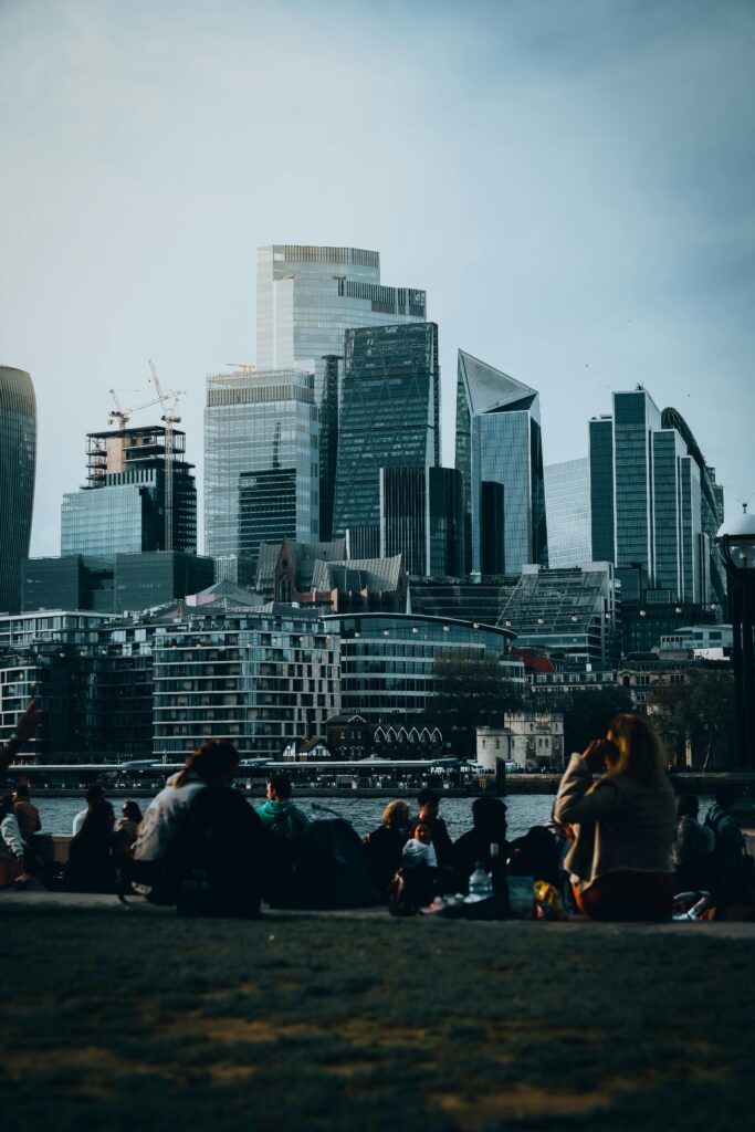 Modern skyscrapers of London overlooking a riverside park with people relaxing on a clear day.