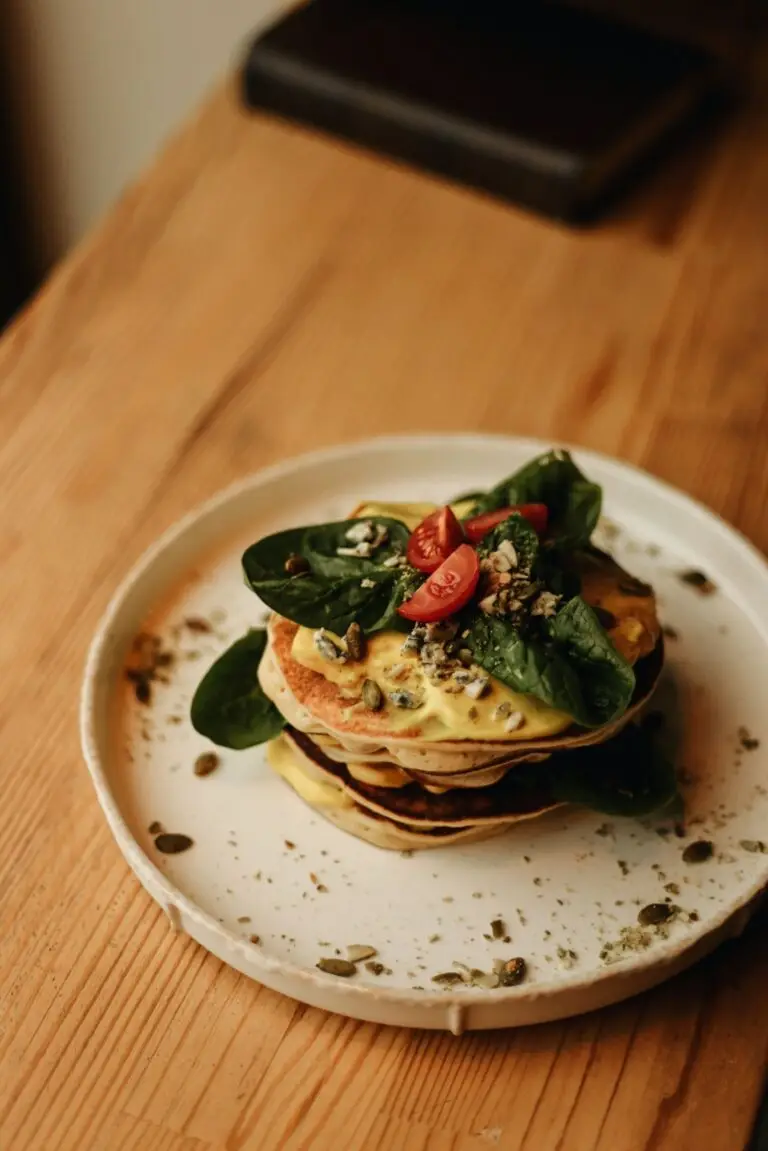 Stacked pancakes with cheese, spinach, cherry tomatoes, and seeds on a wooden table.