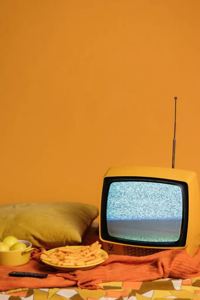 Vintage TV with static on screen surrounded by fries, lemons, and pillow on yellow backdrop.